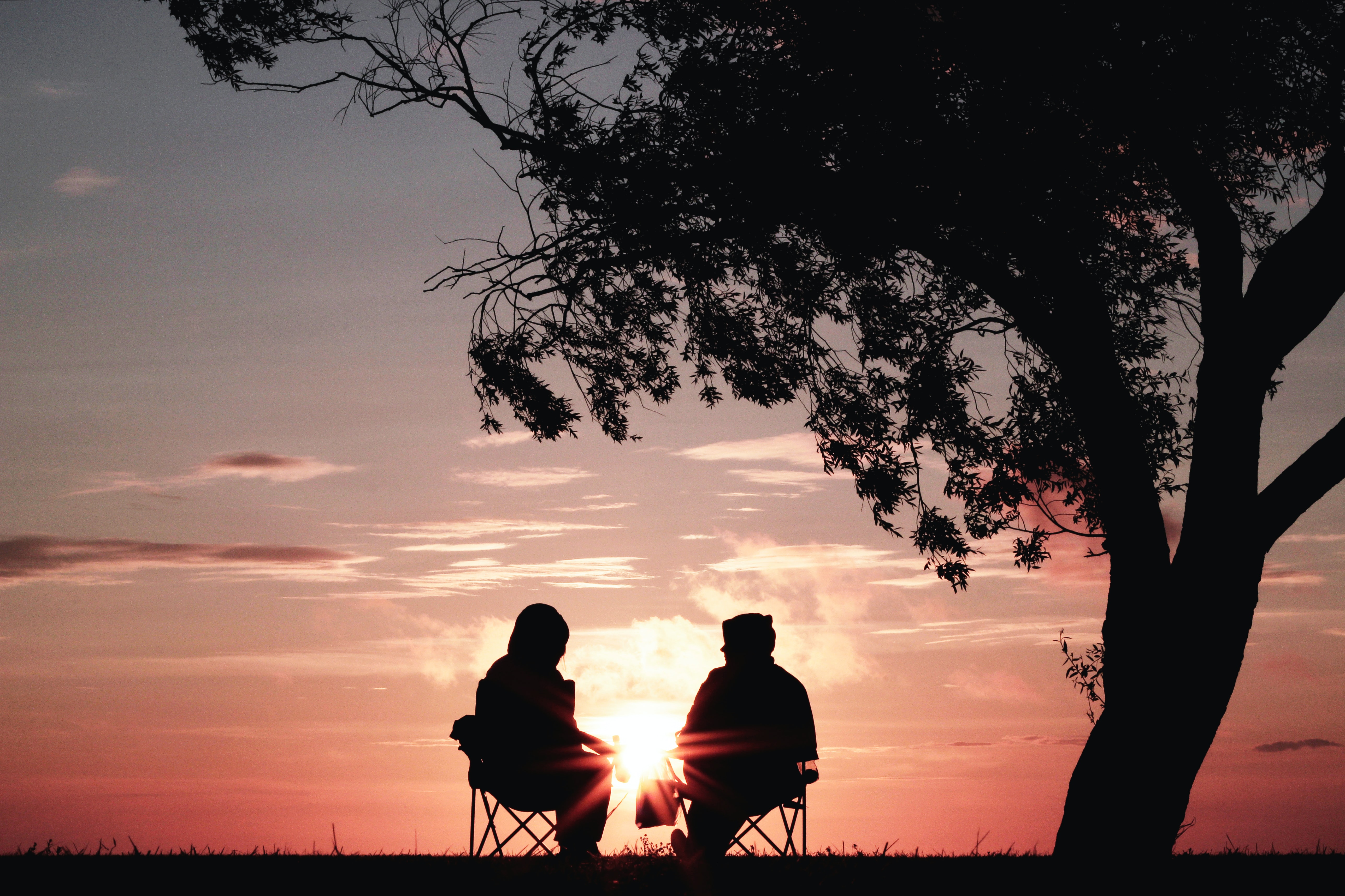 A silouette of two people in the distance sitting on chairs under a tree at sunset having a conversation
